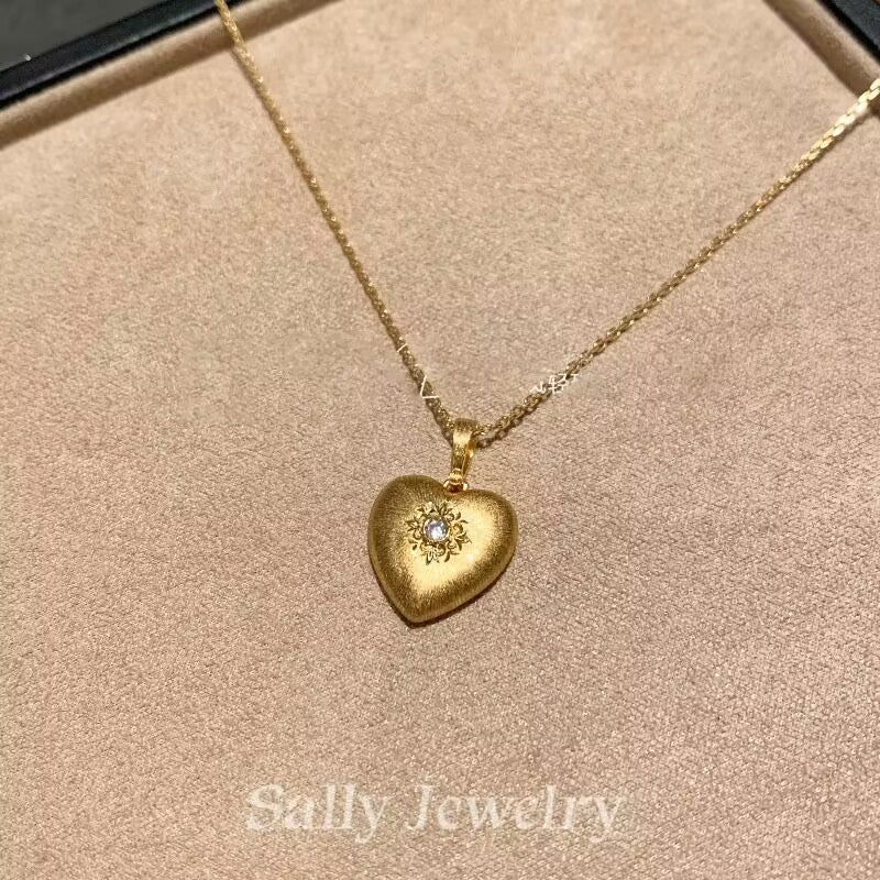 A close-up image of the Heart Brushed Necklace in gold, featuring a heart-shaped pendant with a soft, matte finish. The pendant is suspended from a delicate gold chain, creating a refined and minimalist design. The necklace is displayed against a neutral background, emphasizing the warm gold tone and smooth texture of the heart pendant.