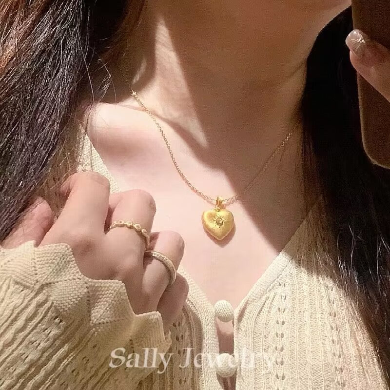 A model wearing the Heart Brushed Necklace in gold. The necklace features a heart-shaped pendant with a brushed, matte finish. The model’s neckline is subtly showcased, with the gold pendant adding an elegant touch to the minimalist design.