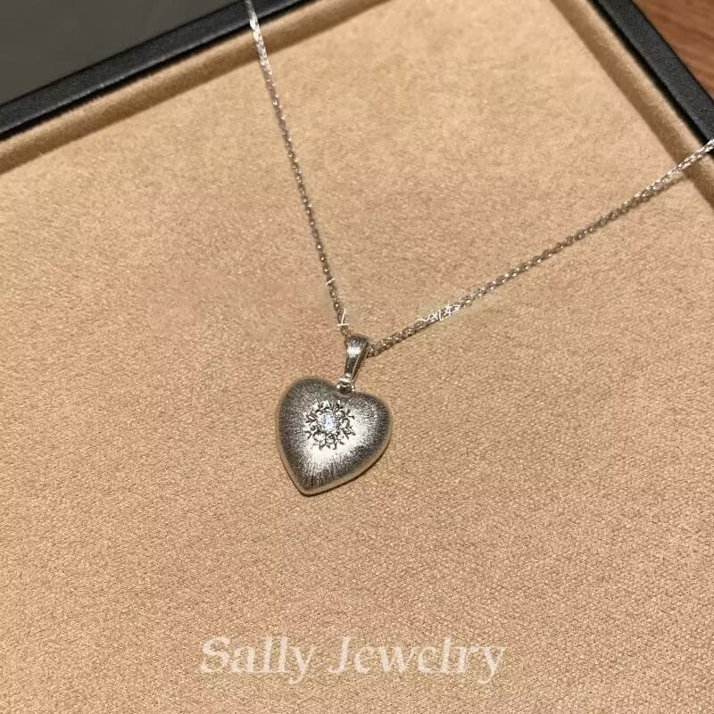 A close-up image of the Heart Brushed Necklace in silver, featuring a heart-shaped pendant with a matte brushed finish. The pendant is suspended from a fine silver chain, highlighting its sleek, minimalist design. The necklace is shown against a simple background, allowing the soft texture and elegant shape of the pendant to stand out.